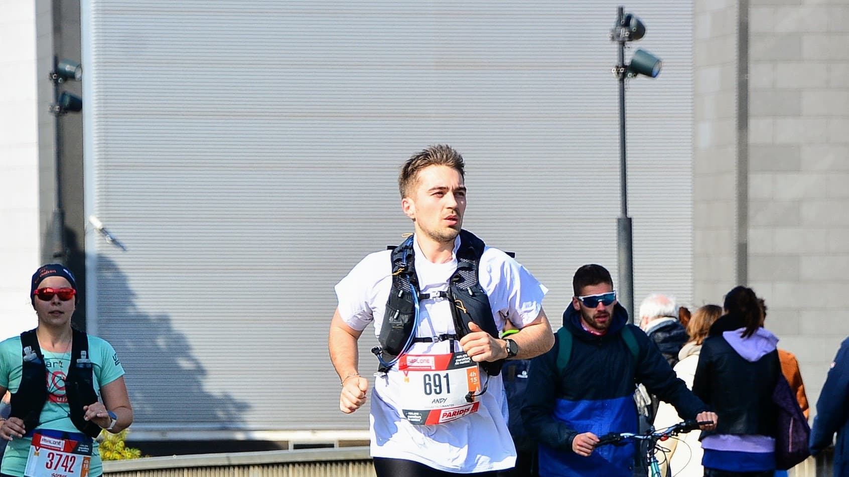 Un coureur participe à une course en plein air, vêtu d'un t-shirt blanc et d'un gilet d'hydratation, avec un dossard numéro 691 bien visible à l'avant. En arrière-plan, une coureuse en t-shirt teal et d'autres personnes exercent diverses activités, notamment un cycliste et des piétons dans un paysage urbain animé, avec des bâtiments gris et un ciel dégagé, reflétant un événement sportif très fréquenté.