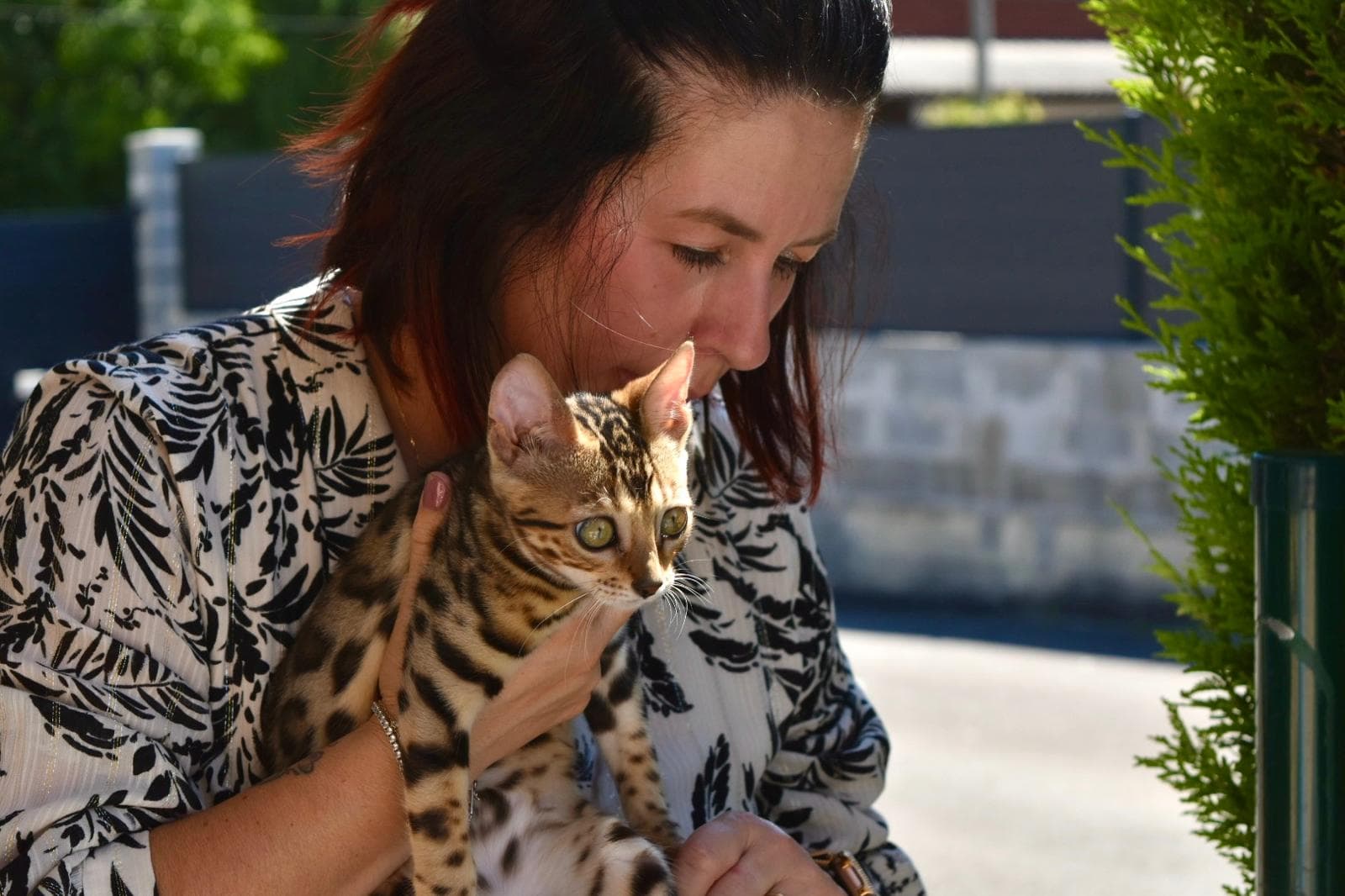 Une femme aux cheveux noirs mi-longs tient doucement un chat Bengal tacheté dans ses bras, regardant le chat avec une expression chaleureuse dans un cadre ensoleillé entouré de verdure.