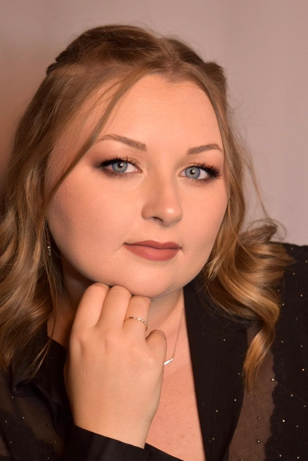 Un portrait rapproché d'une femme aux cheveux blonds ondulés, regardant l'appareil photo avec une expression réfléchie, portant un blouse noire scintillante et un maquillage léger qui met en valeur ses yeux bleus et ses lèvres douces.